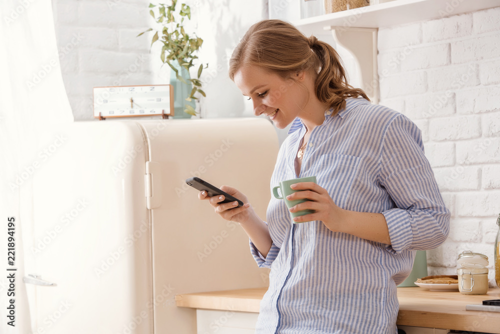 Young woman using smartphone leaning at kitchen table with coffee mug and organizer in a modern home. Smiling woman reading phone message. Brunette happy girl typing a text message. Young woman using smartphone leaning at kitchen table with coffee mug and organizer in a modern home. Smiling woman reading phone message. Brunette happy girl typing a text message.