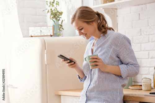 Young woman using smartphone leaning at kitchen table with coffee mug and organizer in a modern home. Smiling woman reading phone message. Brunette happy girl typing a text message.