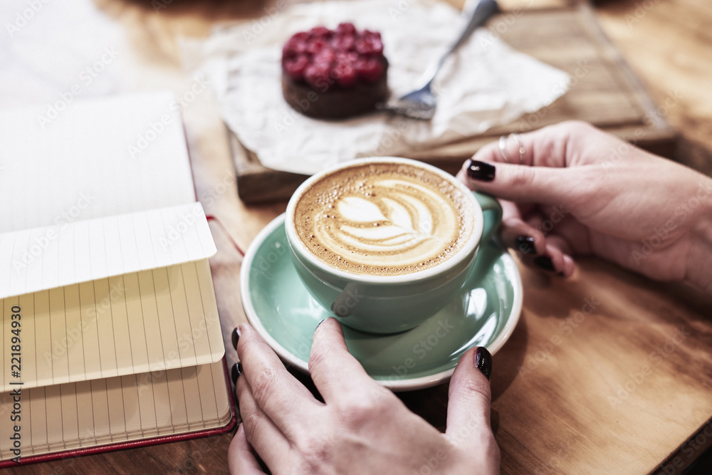 Cup of coffee latte on wooden table or background in woman hands from above. Having lunch in cafe. Opened notebook, space for design template.