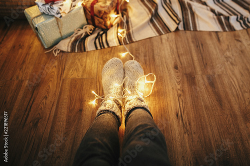 girl legs in stylish warm sock sitting with garland lights at christmas tree with gifts. socks on floor rug in festive room. decor for winter holidays. atmospheric moment