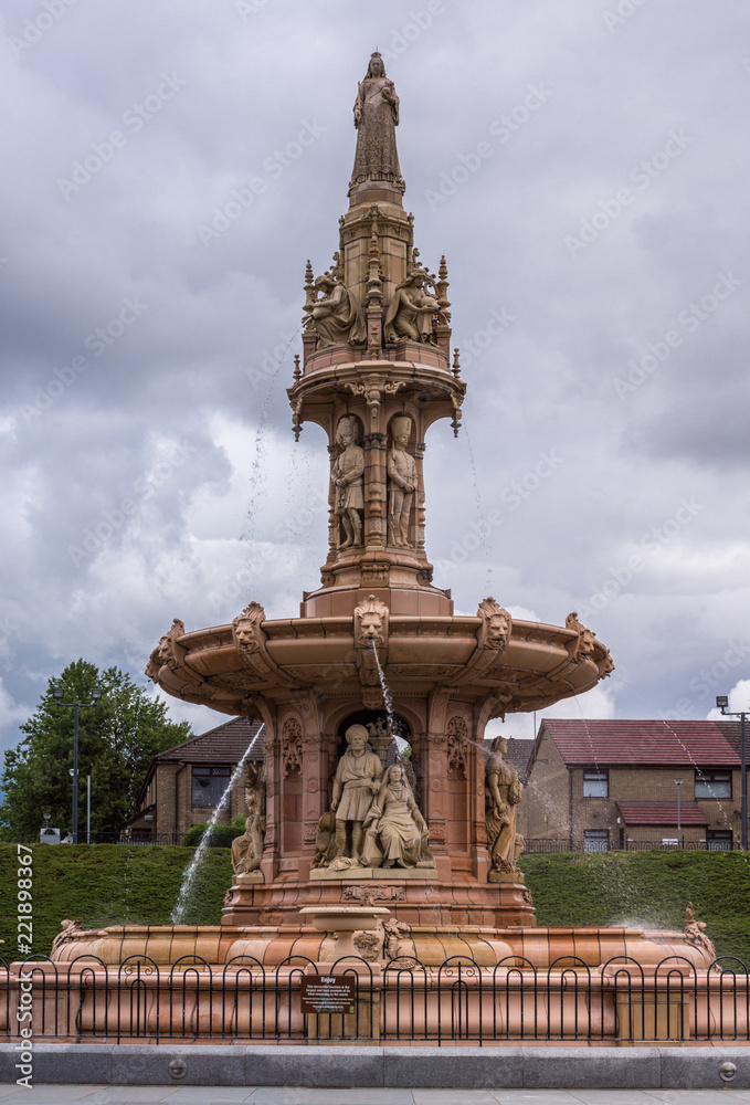 Obraz premium Glasgow, Scotland, UK - June 17, 2012: Doulton Fountain on Glasgow Green under heavy cloudscape. Green foliage and Pendleton building in back.