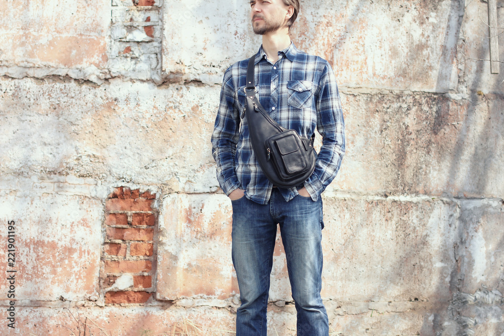 Young hipster man in casual wear with stylish leather crossbody bag standing against concrete wall. Modern city lifestyle