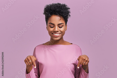 Funny surprised African American female looks with joyful eyes down and indicates as shows something, sees comic things, isolated over lavender wall. Positive amazed young woman poses in studio photo