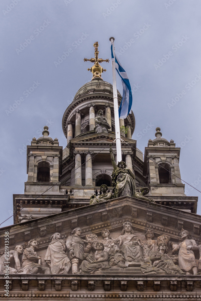 Glasgow, Scotland, UK - June 17, 2012: Closeup of  brown stone fresco behind mesh on facade of City Council building. Scottish flag and top dome of the building with statues.