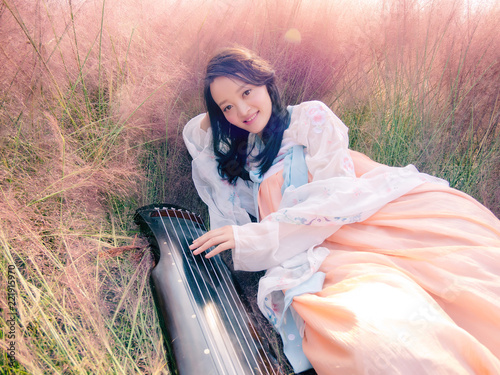 Portrait of beautiful young Chinese woman wearing ancient costume playing guqin in the pink hairawn muhly field. photo