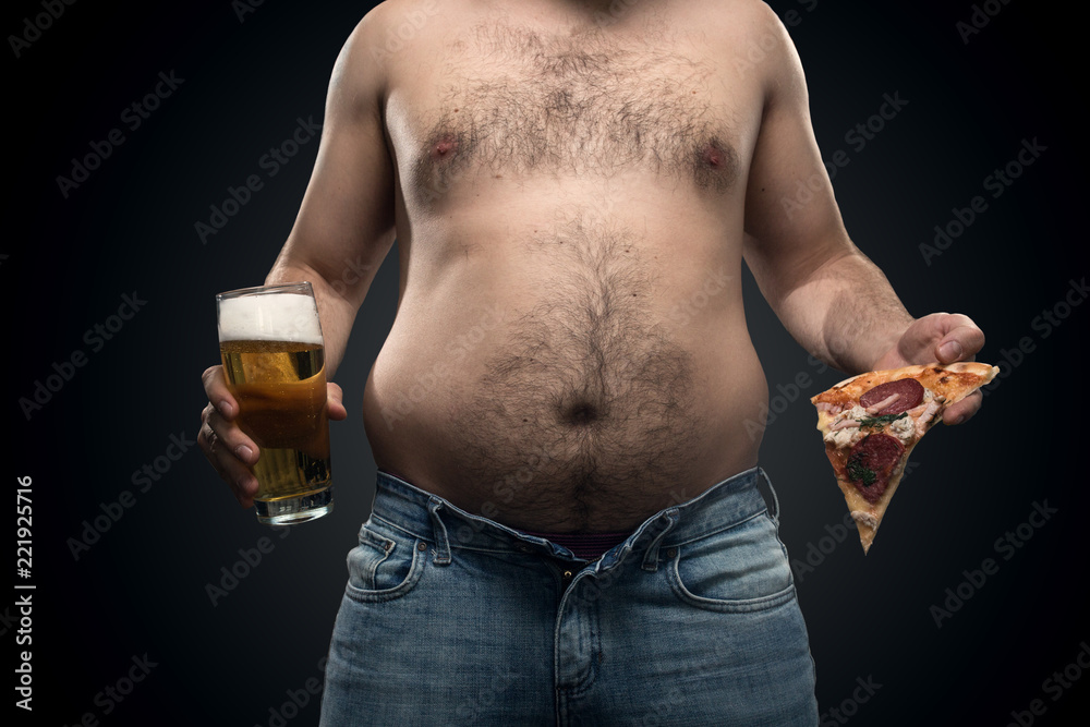 a fat man in jeans with saggy fat on the sides and a hairy belly holding  harmful food-a glass of beer and pizza on a black background Stock Photo |  Adobe Stock