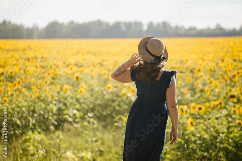 Young beautiful woman in hat stands with her back on a blurred background with a field of sunflowers