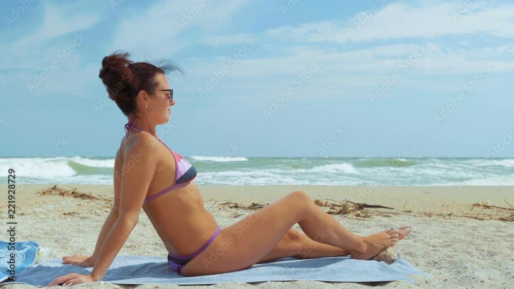 Beautiful Woman in a Swimsuit and Sun Glasses Sits on the Beach and Looks on the Sea Beautiful Woman in a Swimsuit and Sun Glasses Sits on the Beach and Looks on the Sea