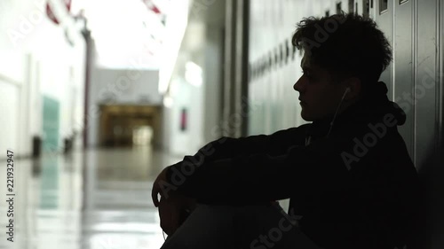 High school teenager sitting in front of his locker alone in empty hallway.