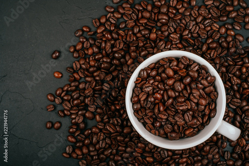 White cup full of with roasted brown coffee beans on the black concrete stone background. Flatlay style.