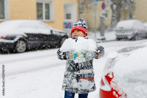 Funny little kid boy in colorful clothes playing outdoors during strong snowfall. Active leisure with children in winter on cold snowy days. Happy child having fun, playing with snow. Winter fashion.