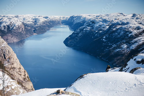 Scenic landscape of the fjord between rocky shore with snow. Top view of the Pulpit Rock, Preikestolen. Man sitting on the big rock. Norwegian mountains. Lysefjord, Norway.