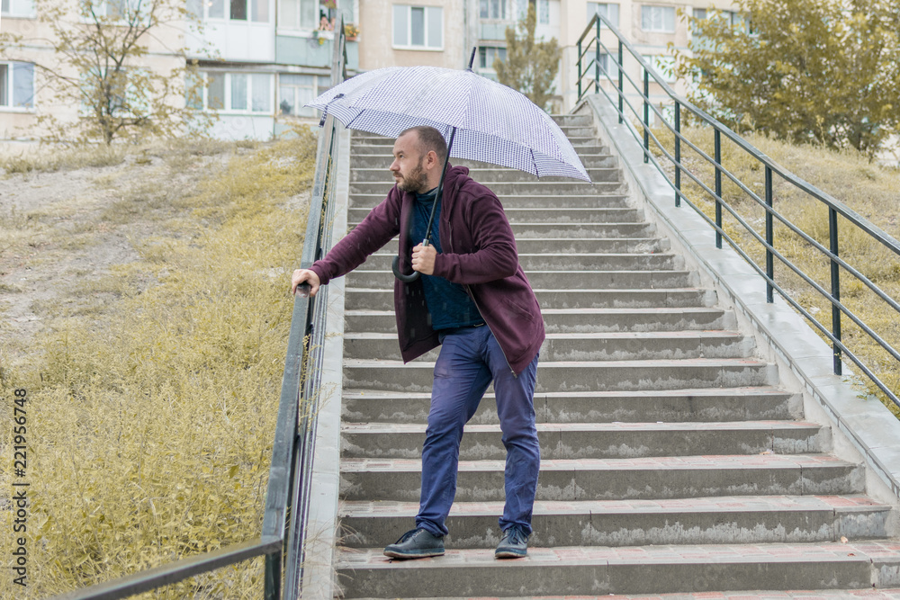 An unshaven man of 35-40 years old in a sweatsuit descends the stairs under an umbrella. An unshaven man of 35-40 years old in a sweatsuit descends the stairs under an umbrella.