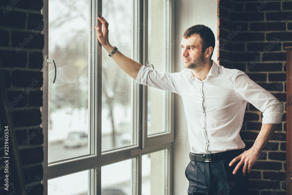 businessman looks through a window from his office. A man in business clothes stands in the room and leans his hand on a window frame