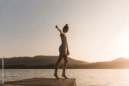 Side view of young caucasian girl dancing on the edge of a dock of a lake in a sunset of a day of summer mountains background photo