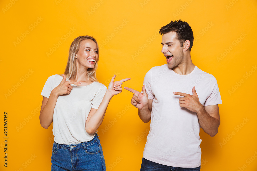 Excited young loving couple standing isolated over yellow wall background pointing to each other.