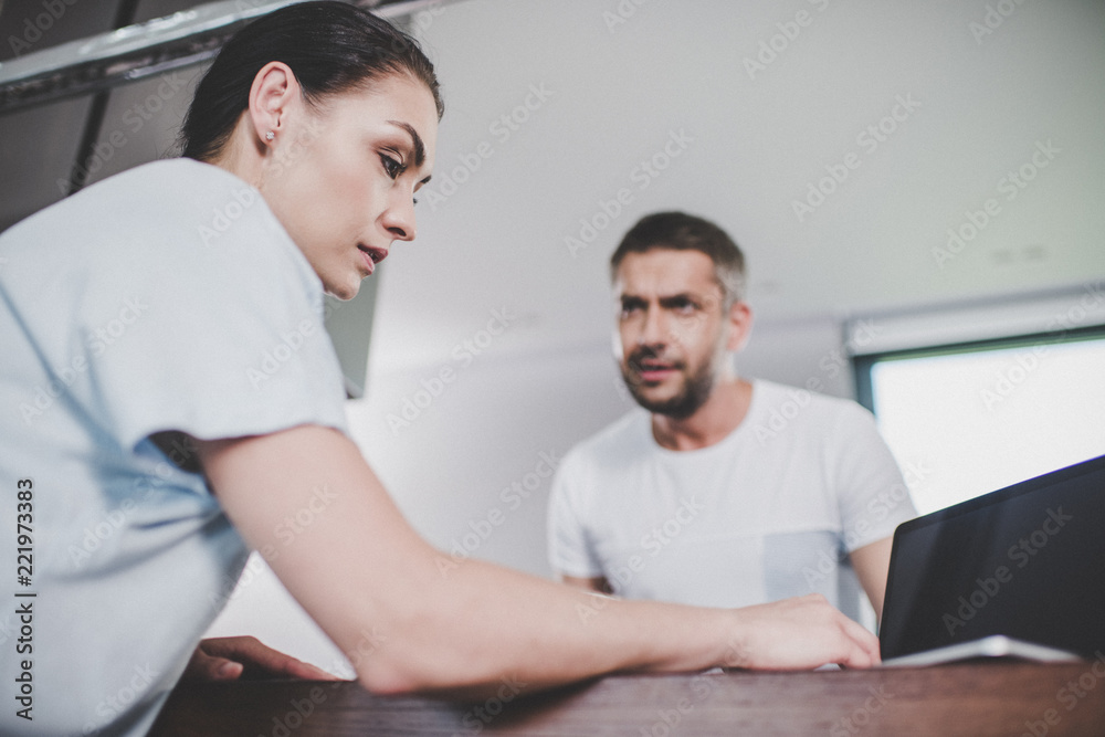 low angle view of girlfriend using laptop, irritated boyfriend looking at her in kitchen low angle view of girlfriend using laptop, irritated boyfriend looking at her in kitchen