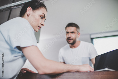 low angle view of girlfriend using laptop, irritated boyfriend looking at her in kitchen
