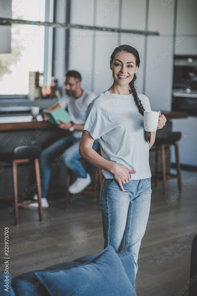 smiling woman with coffee looking at camera while her boyfriend reading book behind in kitchen at home smiling woman with coffee looking at camera while her boyfriend reading book behind in kitchen at home