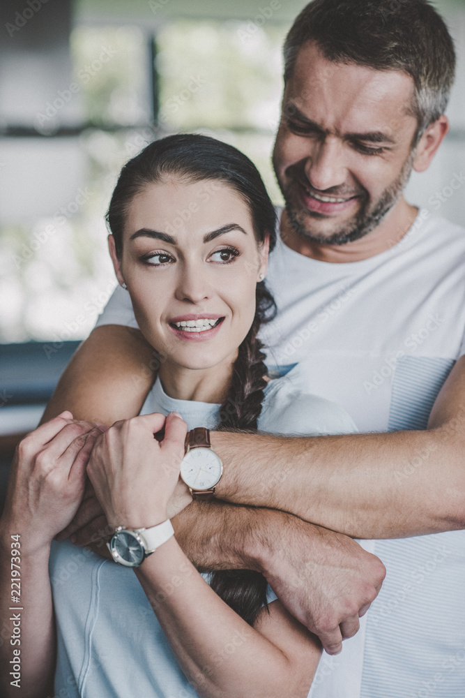 Fototapeta premium portrait of happy man hugging girlfriend from behind in kitchen at home