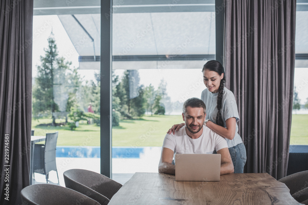 smiling young woman embracing boyfriend while he using laptop at home smiling young woman embracing boyfriend while he using laptop at home