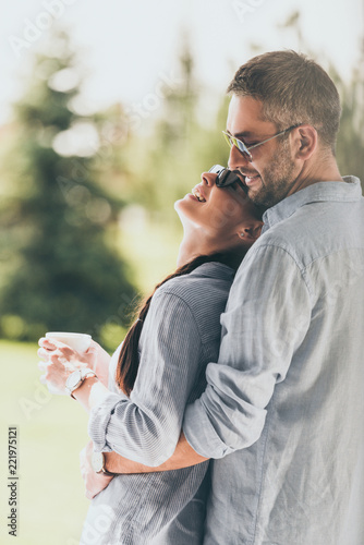 Fotografía side view of happy man in sunglasses embracing girlfriend with coffee cup outdoo