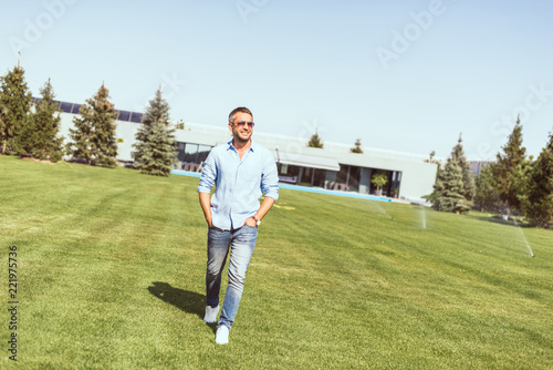 handsome adult man in sunglasses walking with hands in pockets on lawn near country house