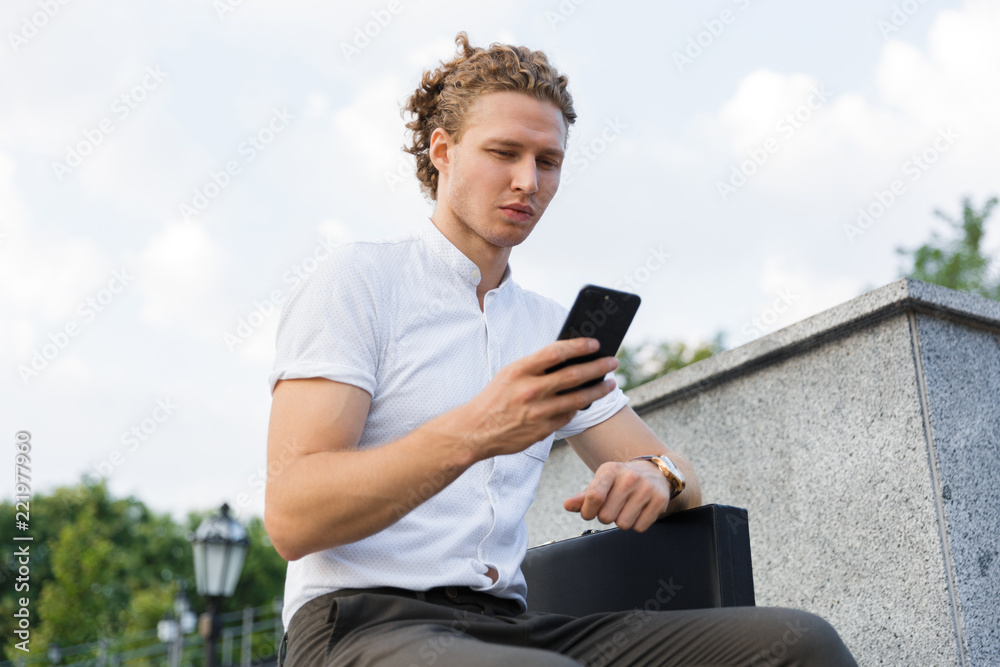 Calm curly business man with briefcase using smartphone