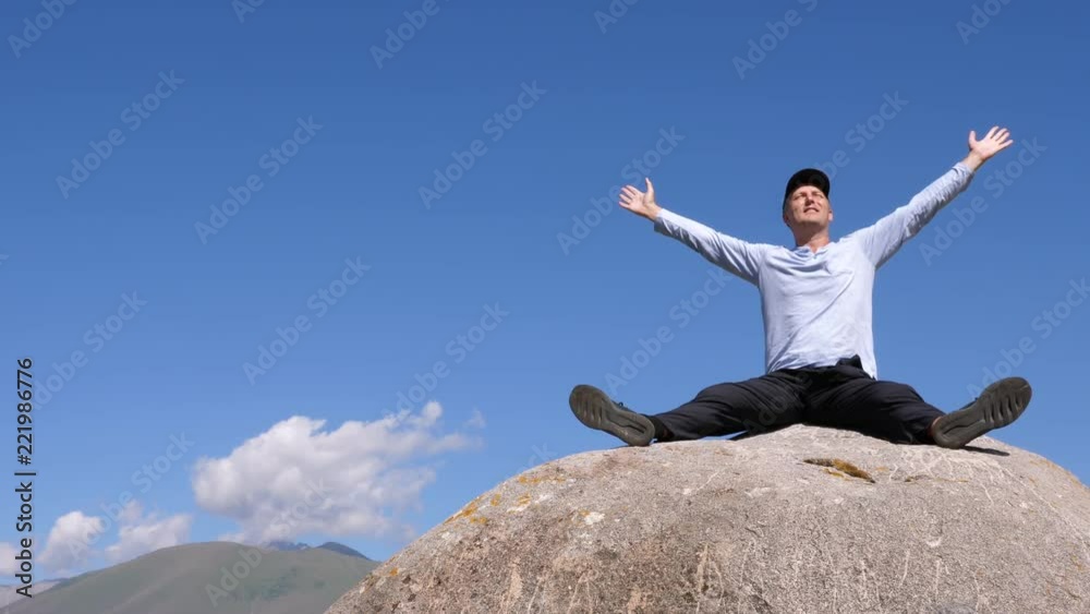 Happy man raising hands up sitting on big stone in mountain valley Happy man raising hands up sitting on big stone in mountain valley