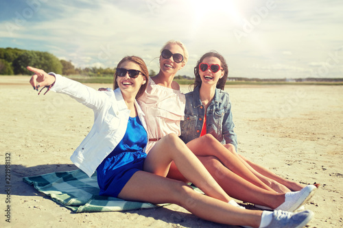 summer vacation, holidays, travel and people concept - group of smiling young women in sunglasses sitting on beach blanket and pointing finger to something