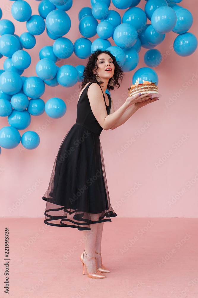 Fototapeta premium young girl holding birthday cake with candles in pink studio room with shiny blue balls decor
