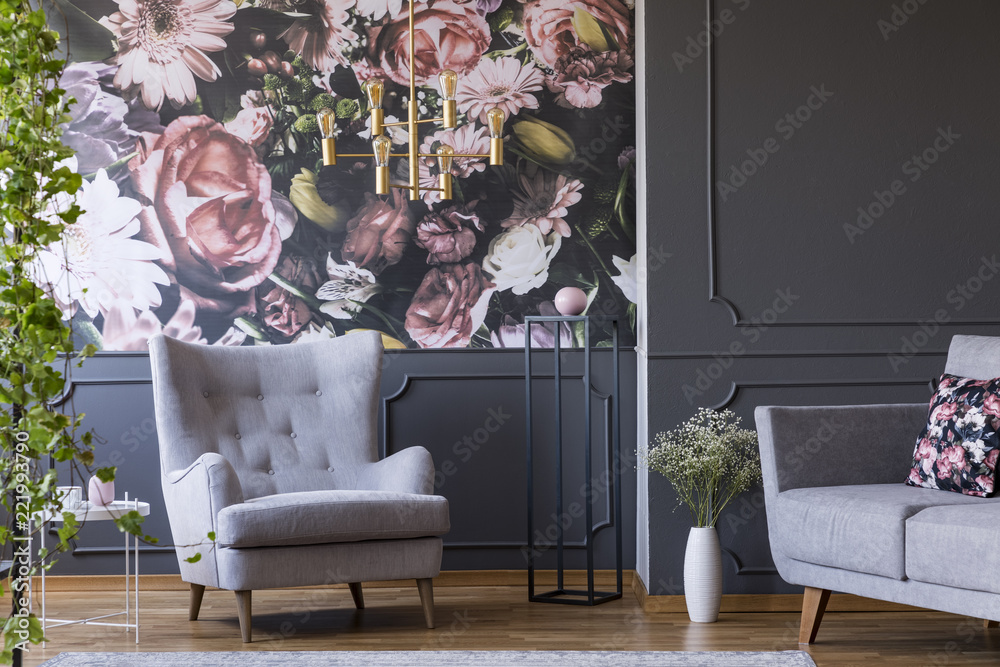 Grey armchair against flowers wallpaper in dark living room interior with sofa and plant. Real photo - obrazy, fototapety, plakaty 