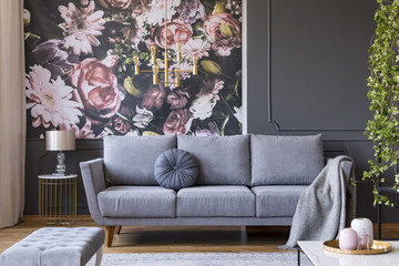 Blanket on grey couch in living room interior with flowers wallpaper and lamp on table. Real photo