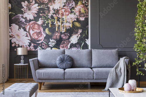 Blanket on grey couch in living room interior with flowers wallpaper and lamp on table. Real photo