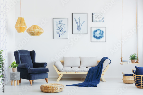 Blue armchair next to a grey sofa, wicker pouf and lamps in a living room interior. Real photo