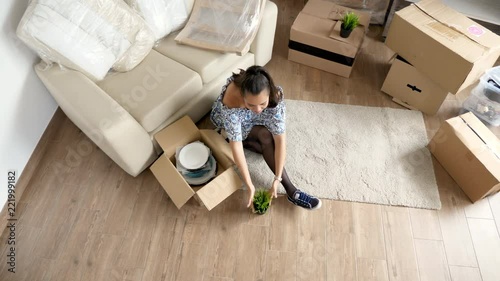 Top view woman unpacking plates and plants from cardboard boxes while the husband is moving furniture in their new house
