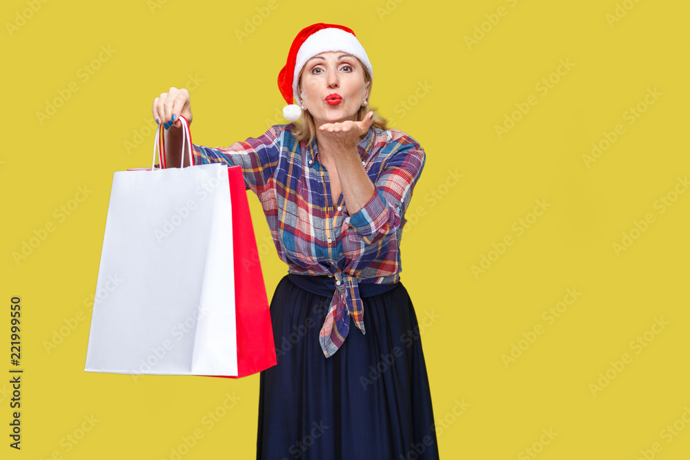 Portrait of kindly beautiful adult woman in red santa cap and checkered shirt standing, holding shopping bags and sending you air kiss, looking at camera. Indoor, studio shot, yellow background