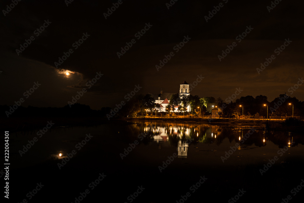 Church in Wieliszew in Poland at night reflecting in the water and with the moon piercing through the clouds