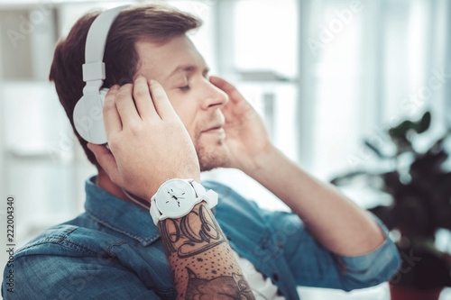 Deep in thoughts. Pleased male person touching headphones while enjoying favorite music