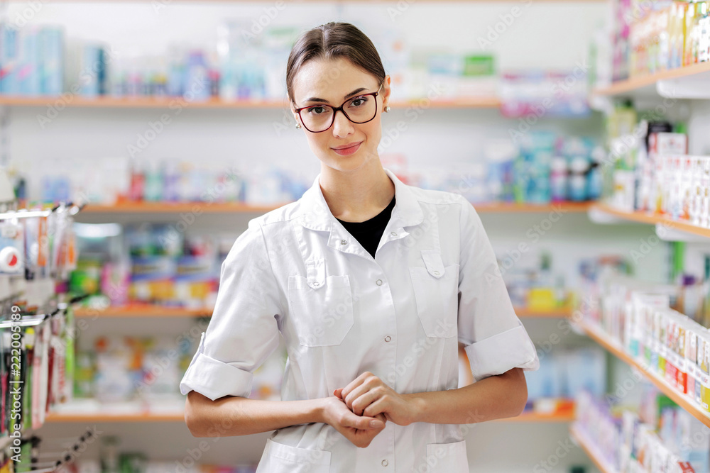 A pretty smiling young lady with dark hair and glasses,wearing a white coat, stands by sjelves in a pharmacy with her arms crossed.