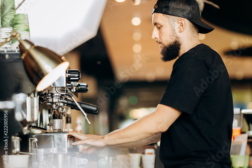 A stylish young man with beard,wearing casual clothes,cooks coffee in a coffee machine in a cozy coffee shop.