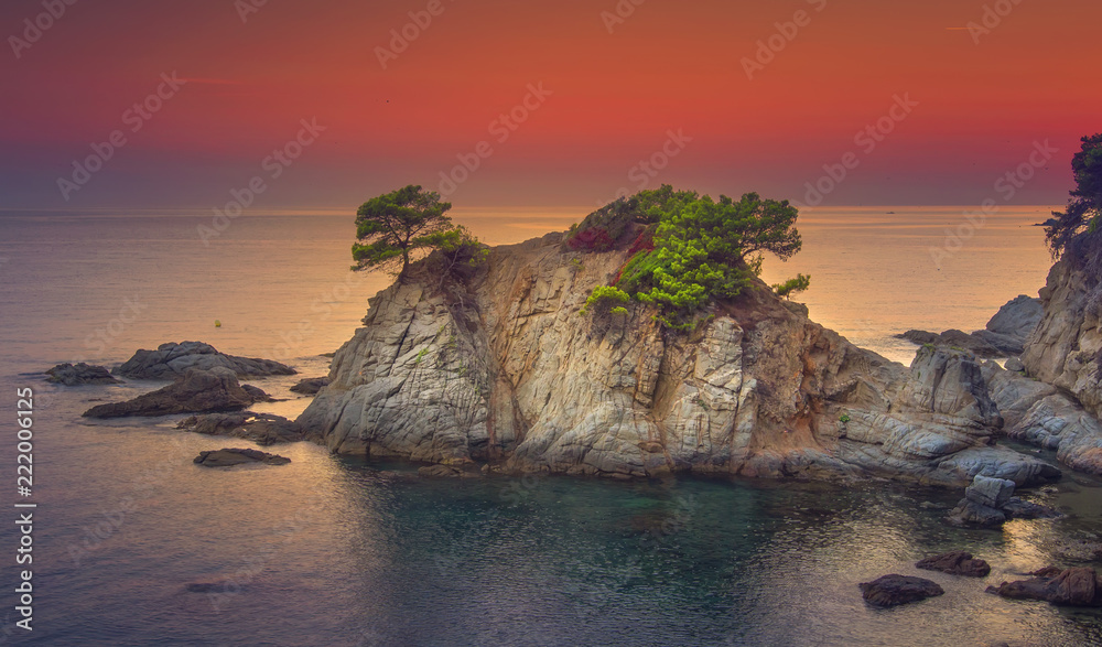 Scenery rocks and mountain on coastline in mediterranean sea at sunrise. Seascape of spanish beach at dawn. Beautiful view on sea with rocky island in morning. Landscape of sea with colorful red sky.