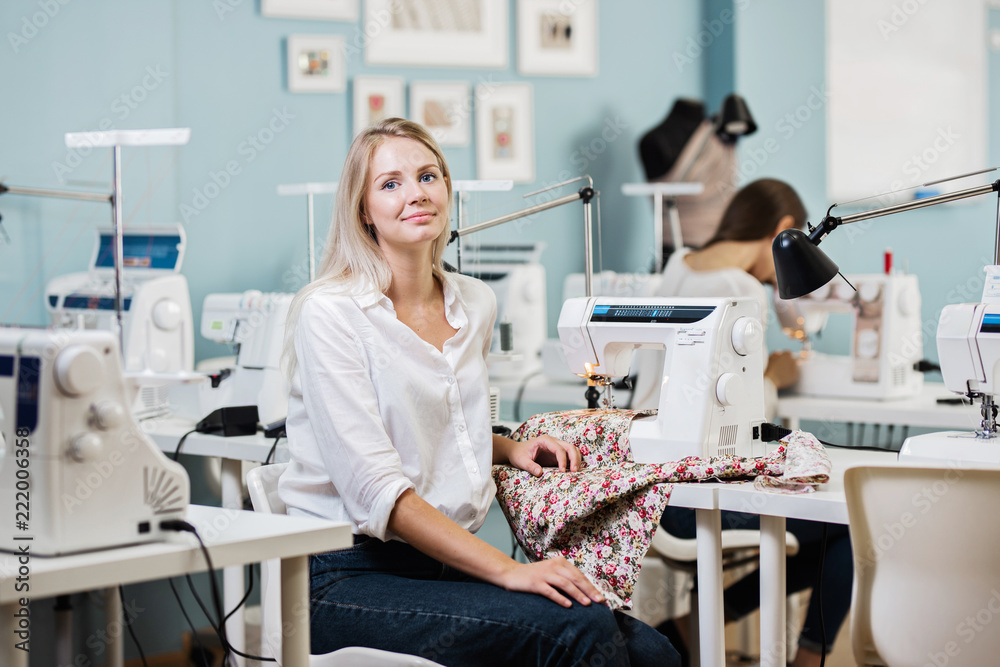A smart-looking pretty blond woman wearing white shirt is sewing with the electric sewing-machine. Fashion, tailor's workshop. A smart-looking pretty blond woman wearing white shirt is sewing with the electric sewing-machine. Fashion, tailor's workshop.