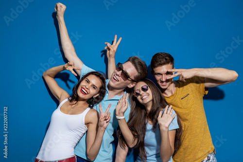 Four good-looking friends are laughing while standing in front of the blue wall having confident and happy looks. Entertainment, having good time. Friendship, relationship.