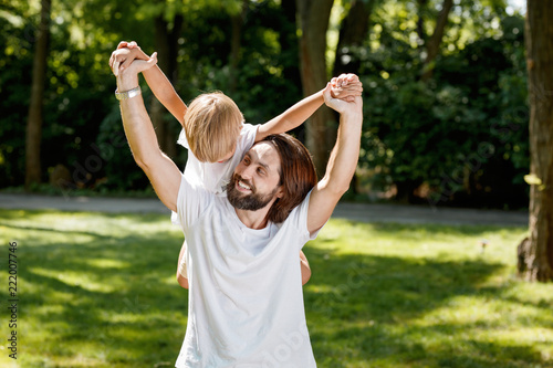 Cheerful handsome man dreesed in white t shirt is holding his little son on his back and laughing outdoor.