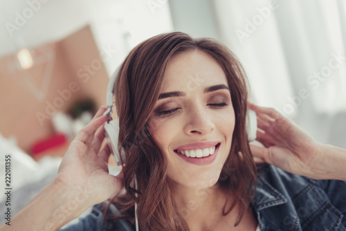 Pleasant moments. Close up of young stylish girl wearing earphones and listening to splendid music while keeping her eyes closed and touching her head
