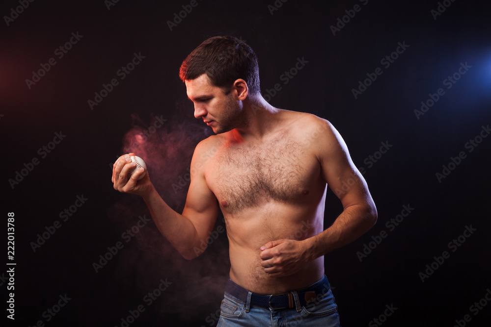 Studio portrait of a muscular sportsman with a baseball player posing on a black background in colored smoke with a baseball in his hand. Concept of training a player in baseball 