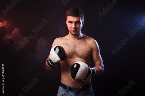Studio portrait of a muscular boxer in professional gloves of European appearance with light bristles and hair on his chest. Smoke in the background is illuminated in blue and red © Тимур Конев