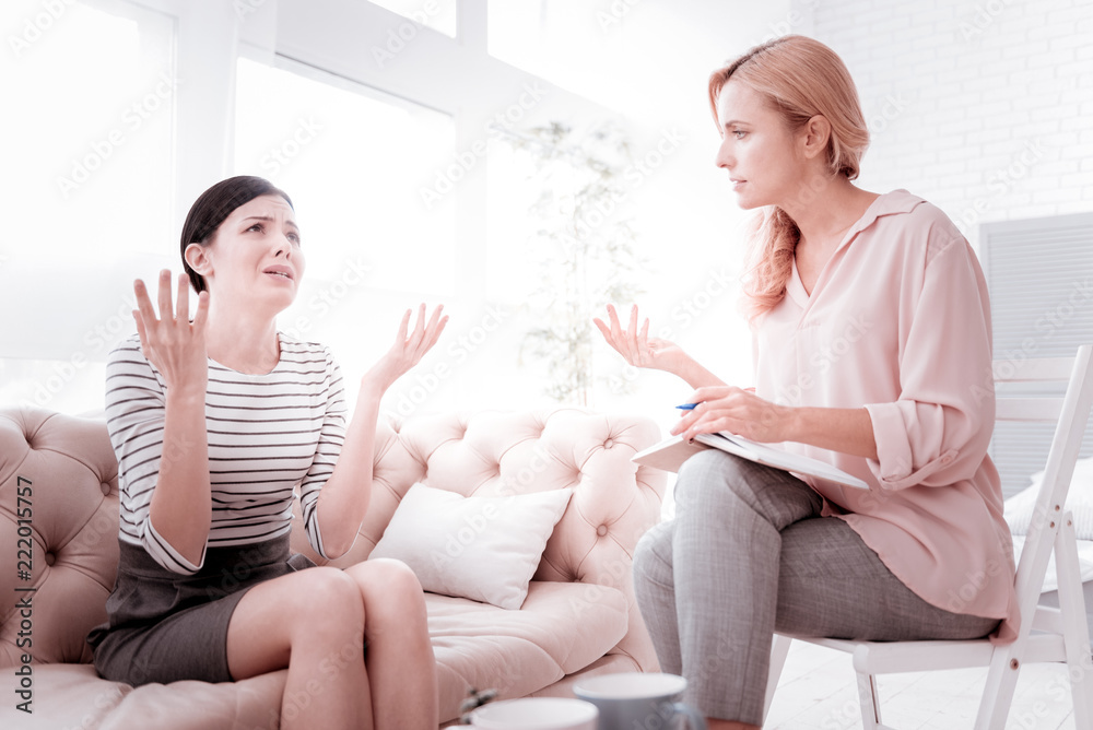 So unfair. Emotional young unhappy woman sitting on a sofa in front of her calm professional psychoanalyst and putting her hands up while crying and talking about a terrible unfair situation at work So unfair. Emotional young unhappy woman sitting on a sofa in front of her calm professional psychoanalyst and putting her hands up while crying and talking about a terrible unfair situation at work