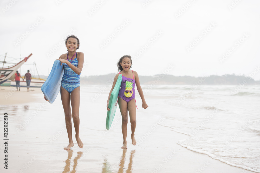 Two beautiful young mixed race sister girls walking on the beach wearing swim wear laughing with boogie boards Two beautiful young mixed race sister girls walking on the beach wearing swim wear laughing with boogie boards